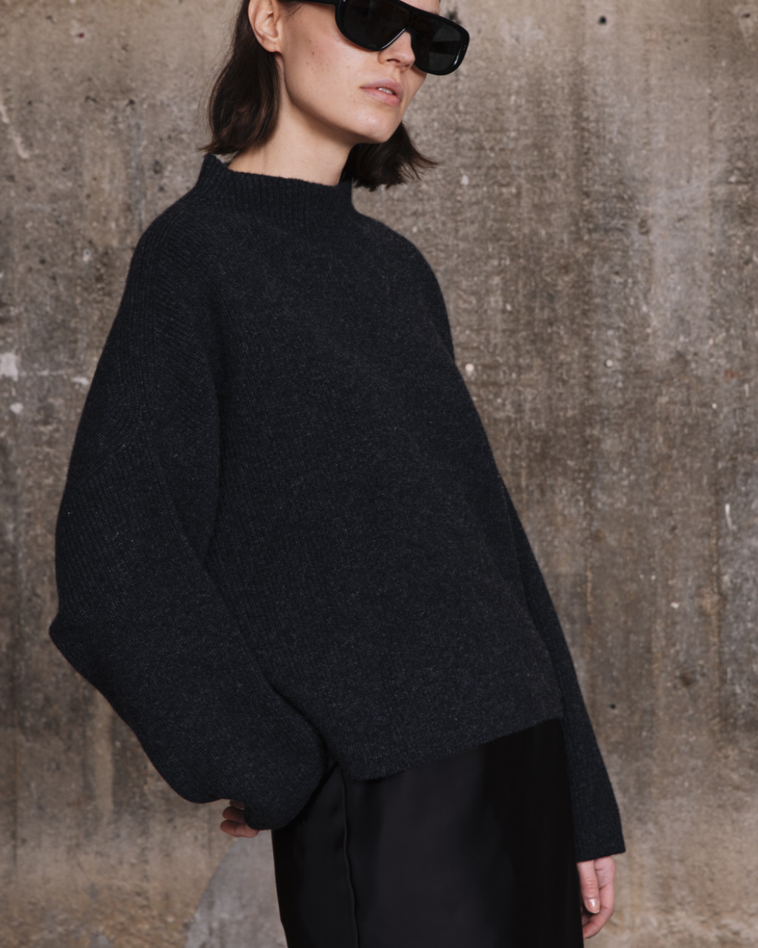 Woman wearing a dark gray sweater and sunglasses against a textured wall.