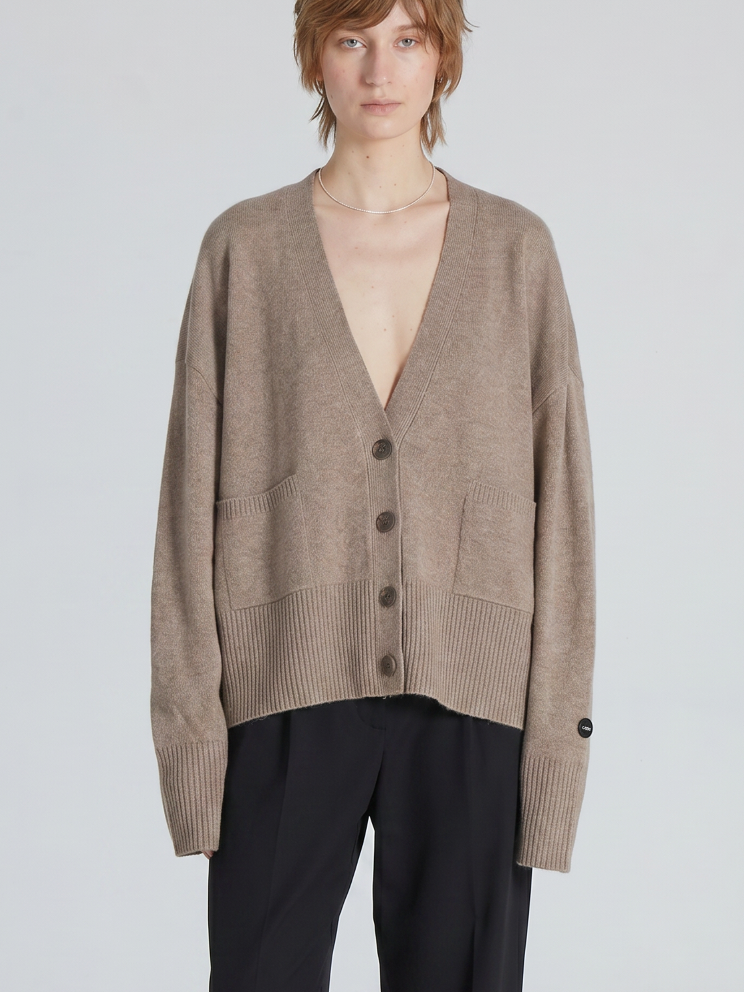 A model wearing a taupe brown knit cardigan with a deep V-neck, front buttons, and front patch pockets, paired with black trousers, against a light gray studio background.