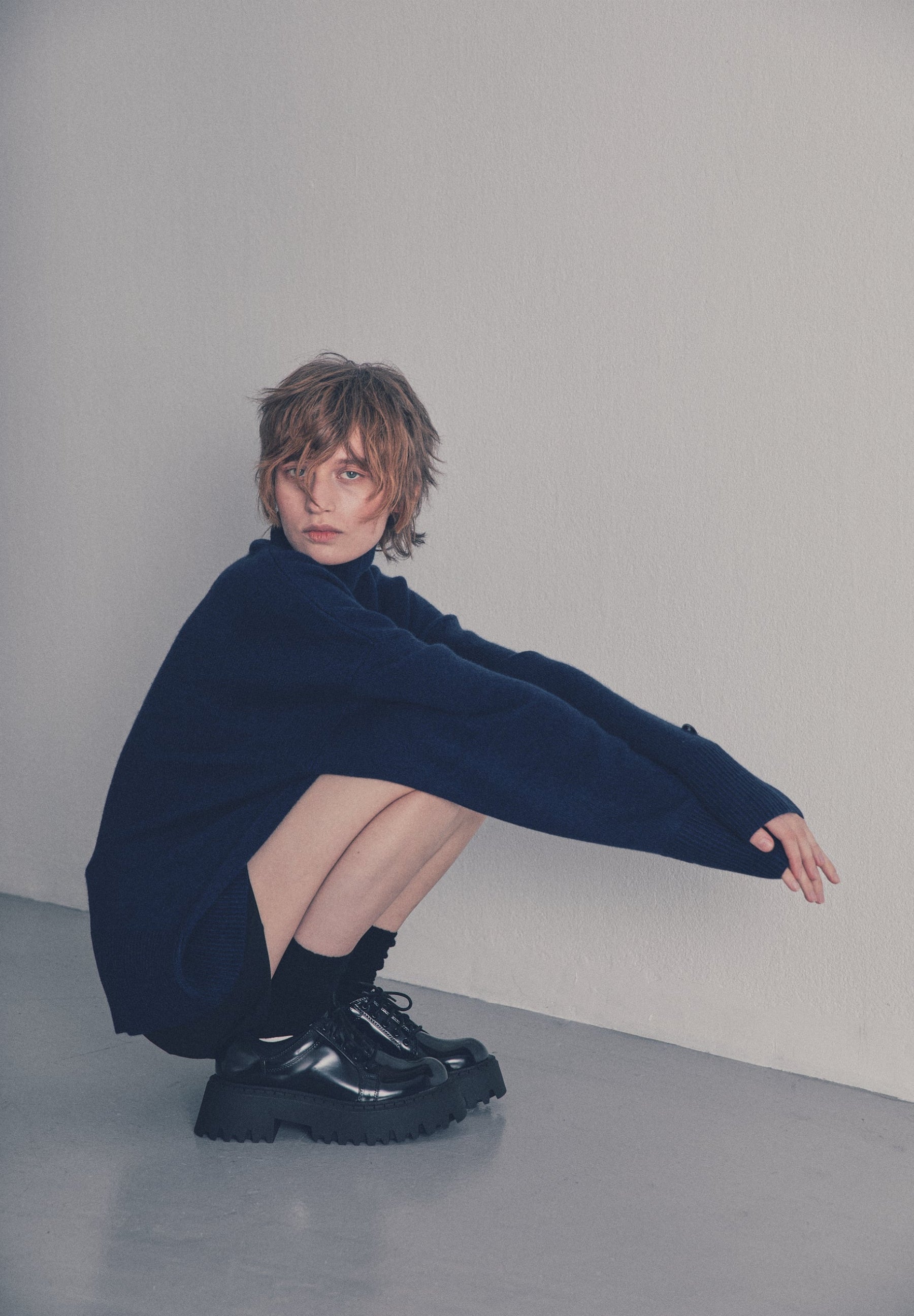 A model with short, layered blonde hair crouches in a minimalist studio setting, wearing an oversized dark navy blue knit sweater, black socks, and chunky black platform lace-up boots against a neutral light gray wall and floor.