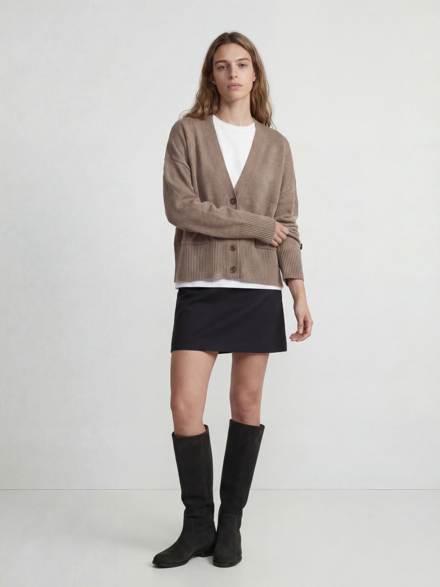 Woman wearing a beige cashmere cardigan, white shirt, black skirt, and black knee-high boots on a white background
