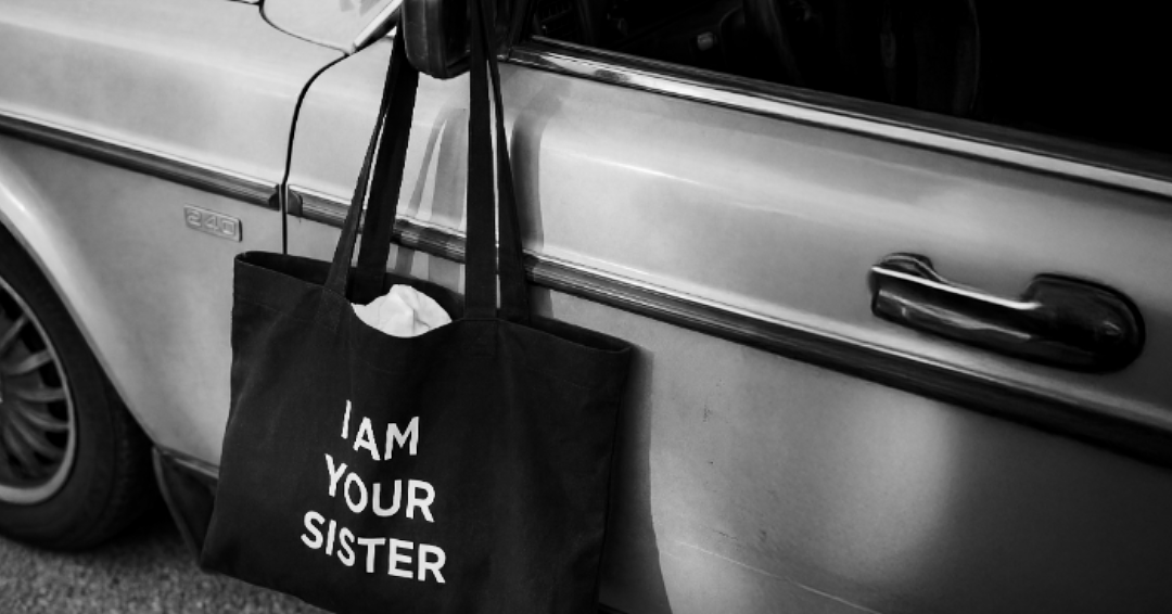Black tote bag with 'I AM YOUR SISTER' text leaning against a car door.