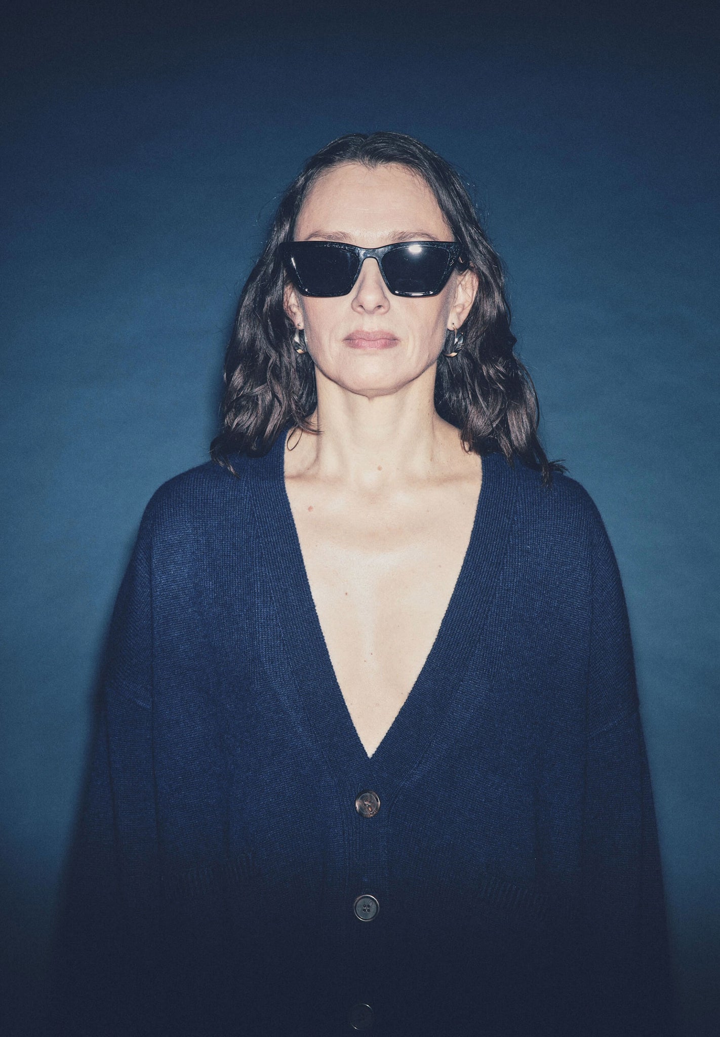 A woman wearing a navy v-neck cardigan with front button-up closure and front pockets, against a dark background.