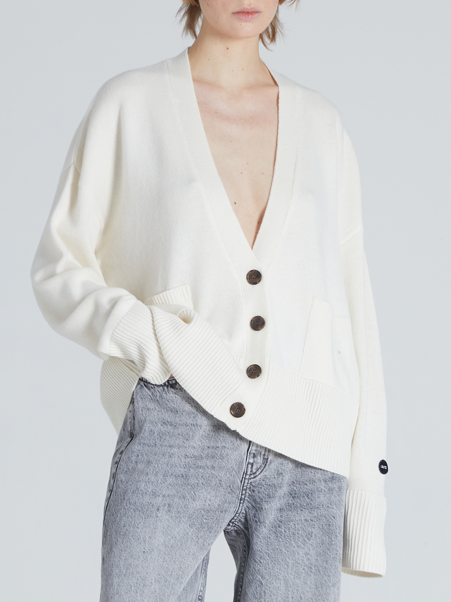 A model wearing an oversized cream-colored knit cardigan with dark brown buttons and light-wash gray jeans, photographed against a solid light gray background.