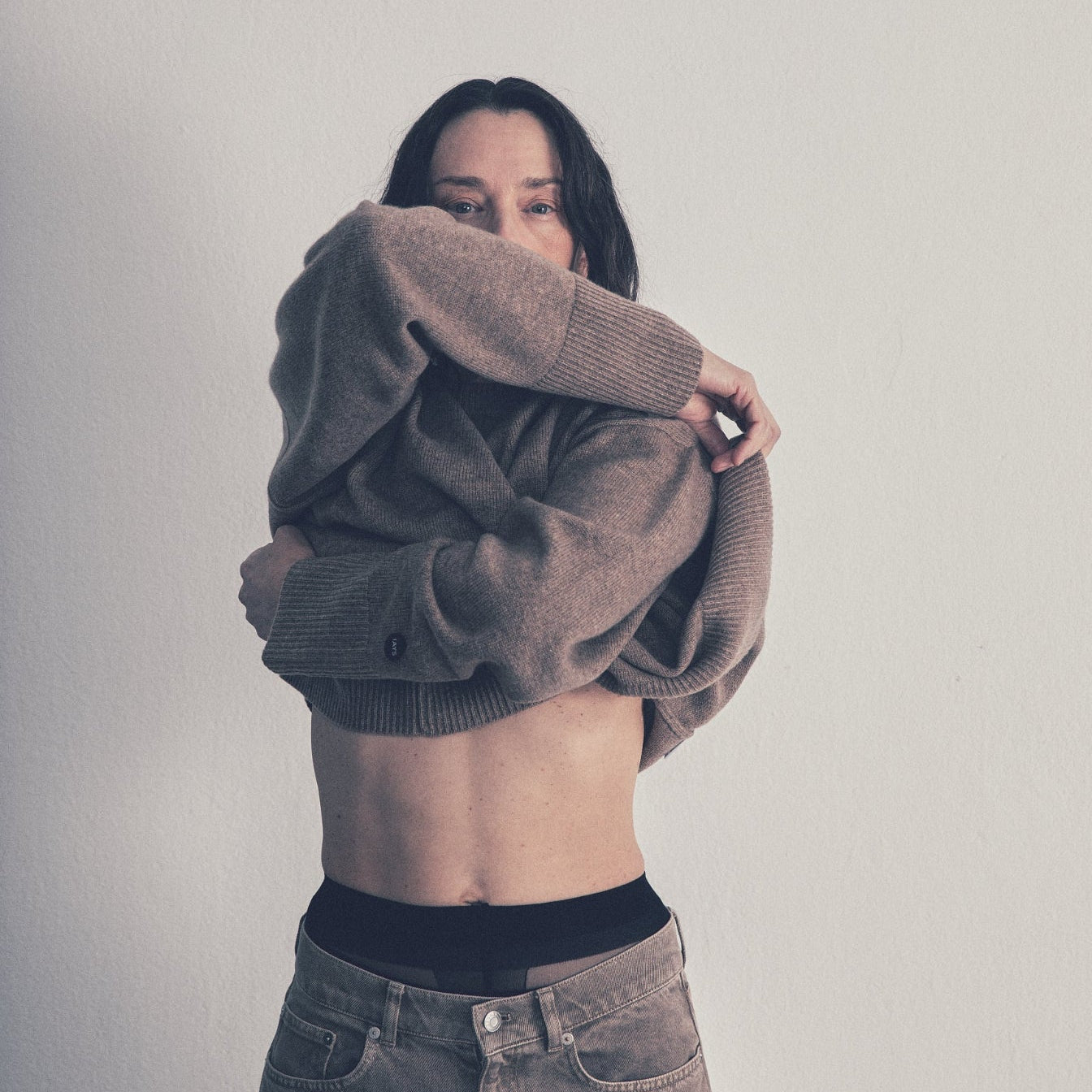 A woman wearing a brown knit cardigan sweater and gray jeans, holding the cardigan up over her head to show its fit and texture against a plain light gray wall.