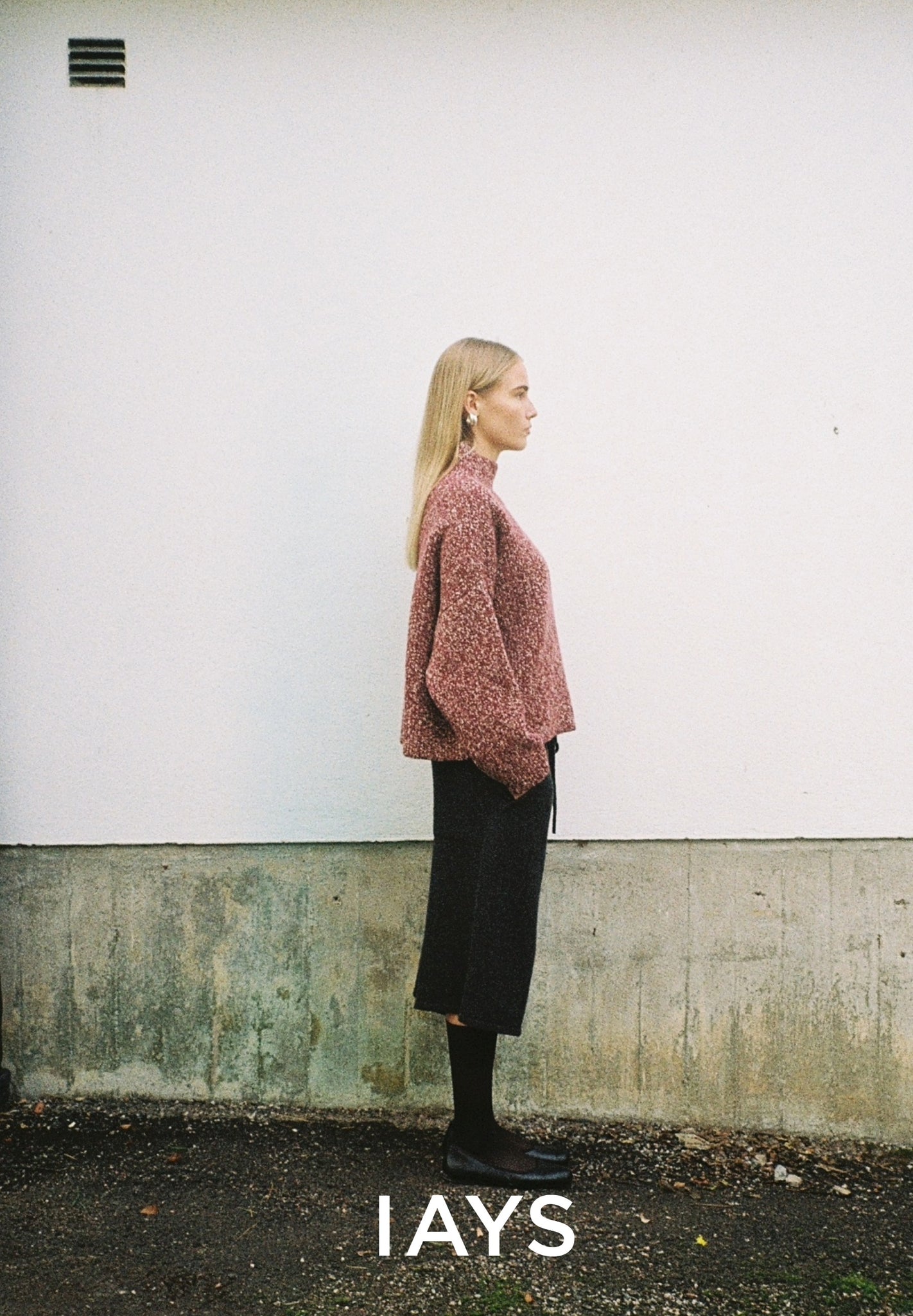 Woman wearing a knitted sweater and black skirt against a white wall with 'IAYS' branding.