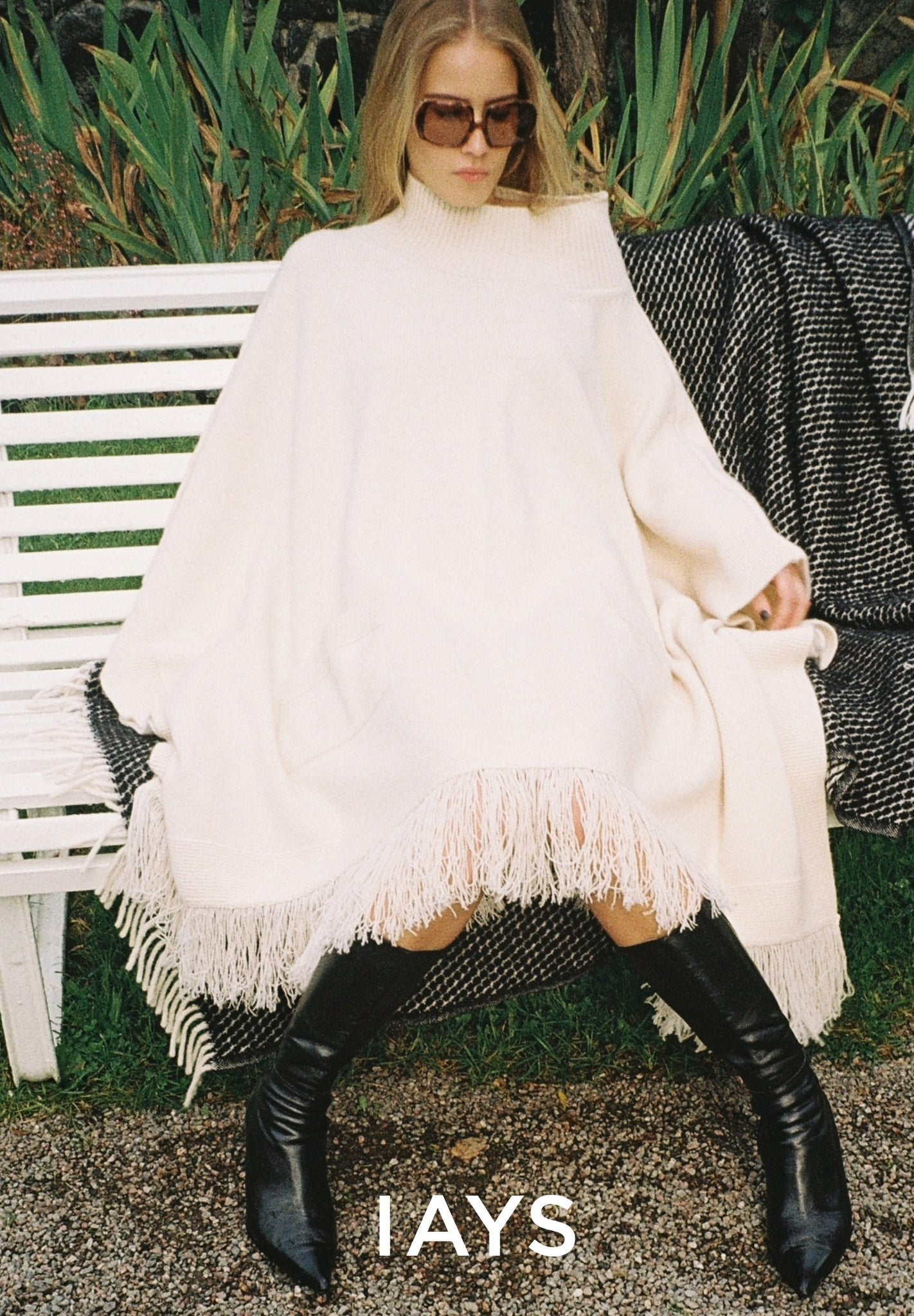 Woman sitting on a bench wearing a white fringe dress and black knee-high boots, with 'IAYS' branding.