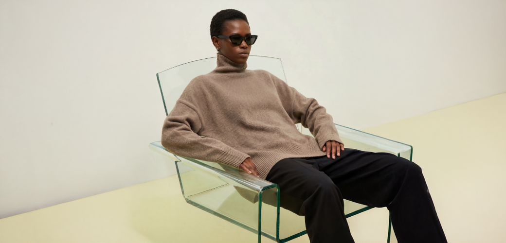 Woman sitting on a chair wearing a beige cashmere sweater and sunglasses in a minimalistic setting.
